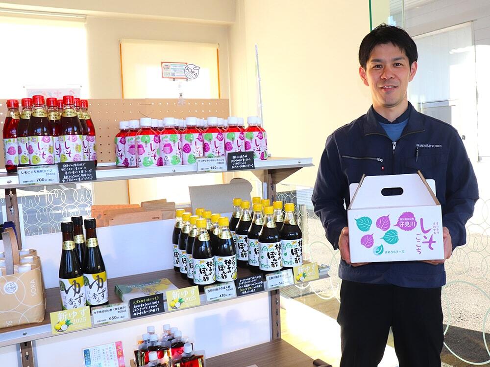 商品棚の横で「すっきりさわやか しそごこち」のギフトボックスを抱えている髙石さんの写真/さめうらフーズ