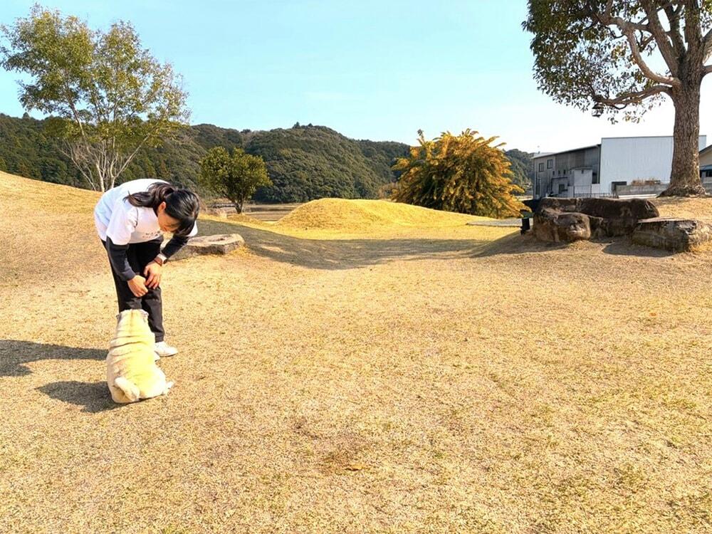 ドッグランエリアを背景に飼い主の近くで座っているパグと飼い主の写真